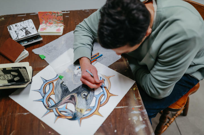 Person seated at a table is drawing a detailed illustration featuring an animal and thorn-like shapes, with colored pencils and sketchbooks nearby.