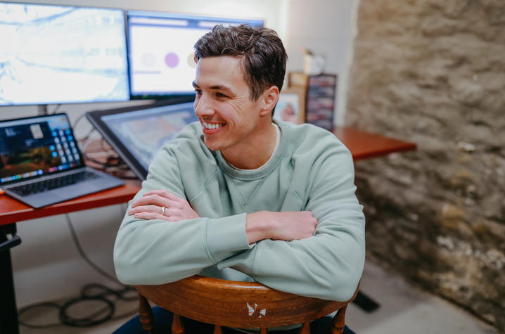 Person in a light green sweatshirt sits on a wooden chair with arms crossed in front of a desk with a laptop and two large monitors; stone wall in the background.