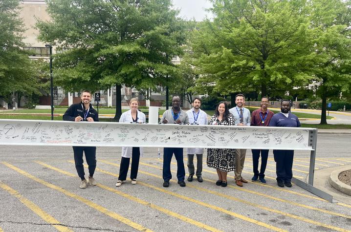 Eight people stand in a line outdoors behind a long, horizontal metal display or sign panel mounted on supports. The group faces the camera and wears a mix of business-casual and professional attire.