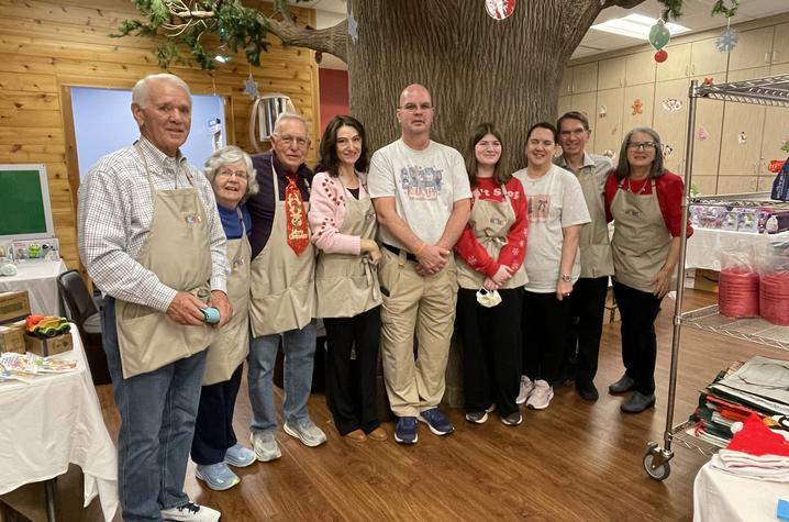 Eight people wearing beige aprons stand together indoors in front of a large artificial tree. The room has wooden walls, tables with toys and gifts.