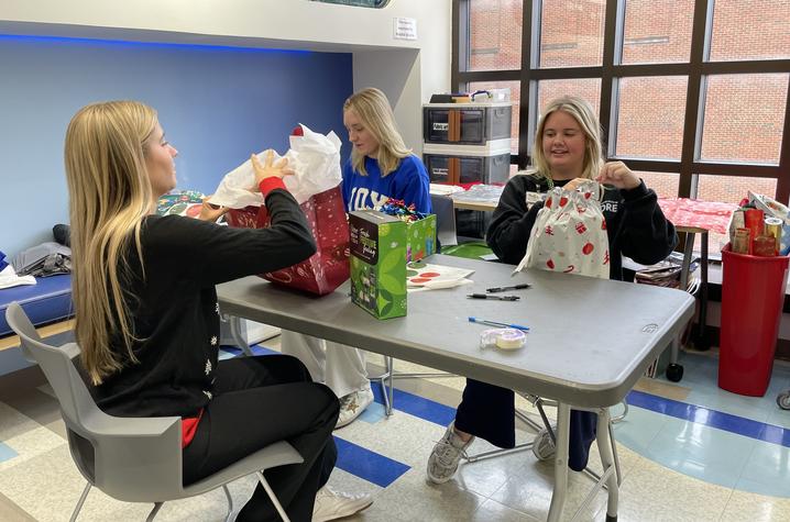 Three people sit at a table wrapping gifts with holiday-themed paper and bags in a bright room with large windows