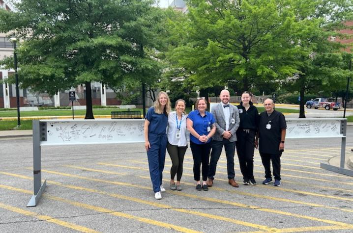 Six people stand together outdoors in a parking lot behind a large horizontal steel beam. The group is arranged in a single row, facing the camera.