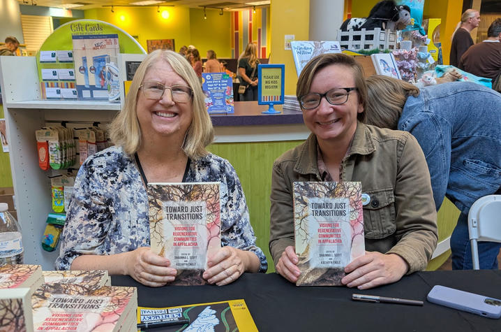 Two people sit at a table at an indoor event, smiling while holding up copies of a book. A display of books and posters is visible behind them.