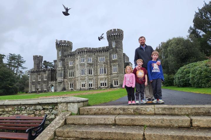 A group of four people stands on stone steps in front of a large gray stone castle with multiple towers and ornate windows. The castle is surrounded by green lawns and trees under an overcast sky. Several birds are flying above the building. 
