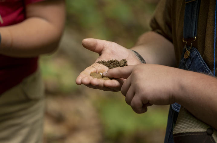 photo of KFLP butterfly experience Close-up of a person outdoors holding a small amount of soil and mud in an open palm while pointing to it with the other hand. Another person stands nearby in the blurred background.