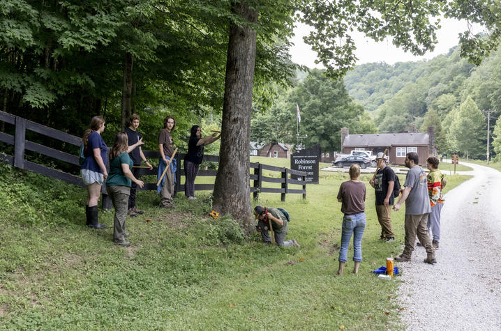 photo of students measuring and identifying trees A group of people gathers around a large tree in a grassy area near the entrance sign for Robinson Forest. Several participants stand along a fence line while others measure or examine the tree, with one person kneeling at the base.