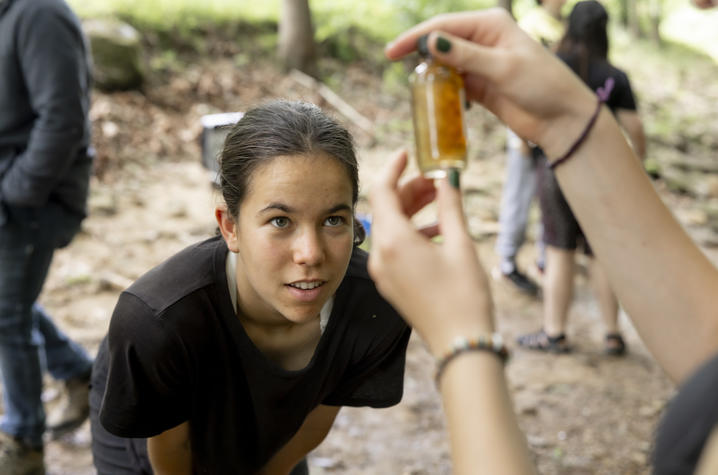 photo of student observing a water sample Close-up of a participant in a wooded outdoor setting examining a small glass vial containing a water sample. Another person holds the vial up to the light while the participant looks closely at its contents. Several other people are visible.