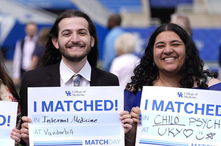 Photo of UK College of Medicine students Two people stand side by side outdoors holding signs that read “I MATCHED!” One sign says “Internal Medicine at Vanderbilt,” and the other says “Child Psych at UK,” with “MATCH” printed along the bottom of each sign.