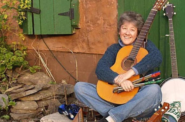 photo of LIza DiSavino leaning at door of house hugging her guitar surrounded by instruments