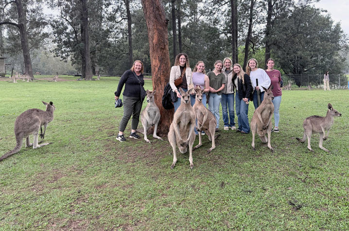 A group of eight people stands on a grassy field in a wildlife park, posing near several kangaroos gathered around a large tree. Additional kangaroos can be seen grazing and resting throughout the open area, which is surrounded by tall eucalyptus trees.