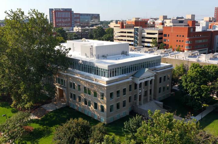 Aerial view of a multi-story academic building surrounded by trees, with additional campus buildings visible in the background.