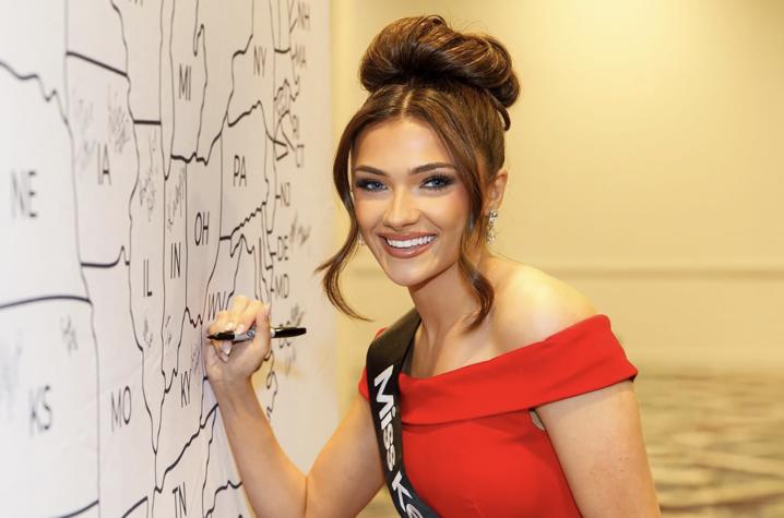 Person in a red off-the-shoulder dress signs a large U.S. map display with a marker while standing beside the states of Ohio, Indiana and Kentucky.