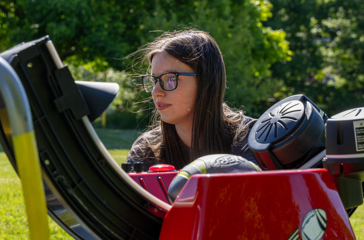 Close-up of a person sitting next to a red adaptive football launcher outdoors. The person is wearing glasses and looking toward equipment on the device. Trees and greenery are visible in the background.