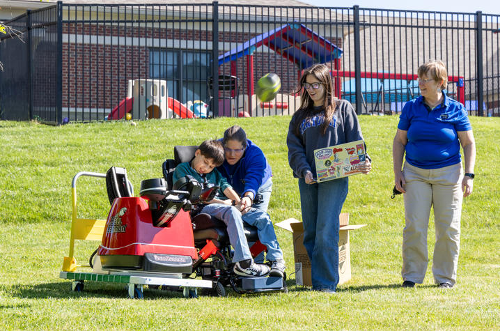 A child operates a football launcher device with assistance from another person during an outdoor activity on a grassy field. Two additional adults stand nearby, and a playground and fence are visible in the background.