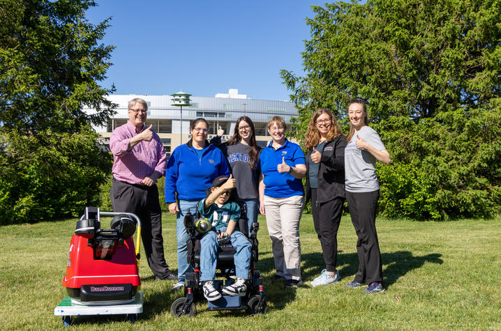 Group photo outdoors featuring a child seated in a motorized wheelchair surrounded by six adults. A red adaptive football launcher is parked beside the group, with trees and a university building visible in the background.