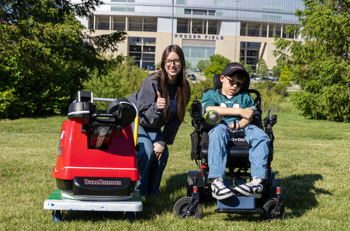 A child seated in a powered wheelchair sits beside a red adaptive football launcher on a grassy lawn. Another person kneels next to the child. A campus building and trees are visible in the background.