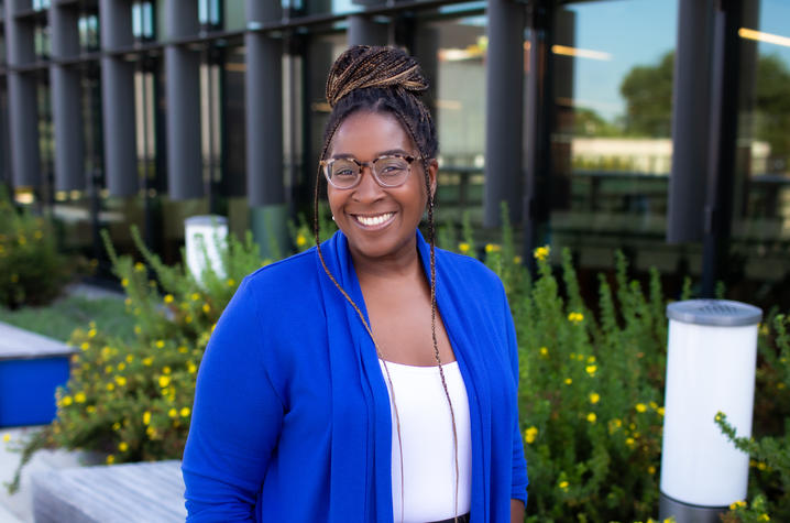 A smiling person wearing glasses and a bright blue cardigan stands outside a modern building surrounded by greenery and yellow flowers.