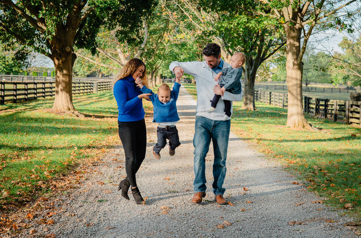 Two adults walking on a tree-lined gravel path, holding two young children, with wooden fences and green grass on both sides.