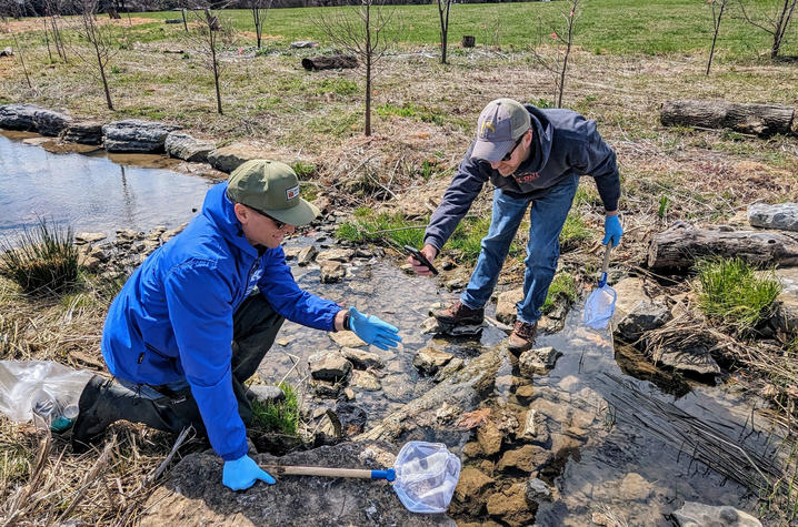 Two people wearing outdoor clothing kneel beside a small stream in a field while collecting water samples.