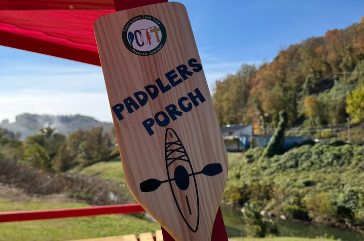 A wooden paddle sign reading “Paddlers Porch” is mounted to a bright red outdoor pavilion overlooking a creek. A walking path, wooded hillside and clear blue sky are visible in the background.
