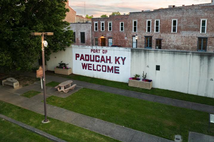 a mural on a white wall that says “Paducah, KY Welcome.” A small grass field and a picnic table are sitting in front of the sign.