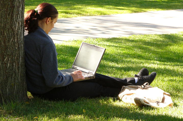 Woman leaning on a tree working on her computer. 