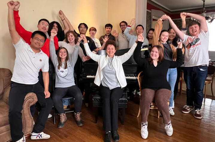 Several college students and a professor gathered around a piano with their hands raised.