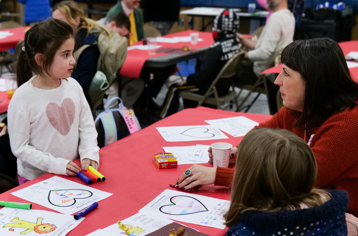 A young girl talks with a woman at a table covered in Valentine-themed coloring sheets and markers, while others sit and chat in the background.