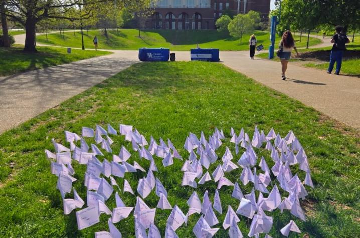 Paper flags arranged in a heart shape on a grassy campus lawn, with a large academic building and people walking on pathways in the background.