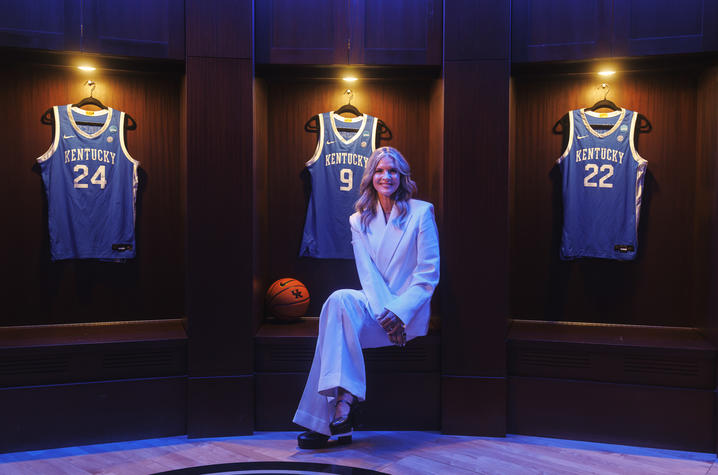 A person dressed in a light-colored suit sits in the center of a display featuring framed University of Kentucky basketball jerseys. Three blue jerseys, including numbers 24, 3 and 22, are mounted in illuminated wooden display cases behind the person.