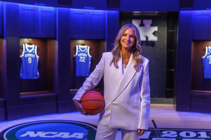 A person wearing a light-colored pantsuit stands in a display space featuring illuminated University of Kentucky basketball jerseys mounted in wooden cases. The person holds a basketball while standing on a floor marked with NCAA tournament branding.