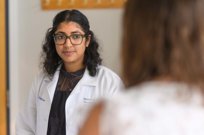 Person wearing glasses and a white lab coat looks toward another person during a conversation. The second person appears out of focus in the foreground, with an indoor office setting in the background.