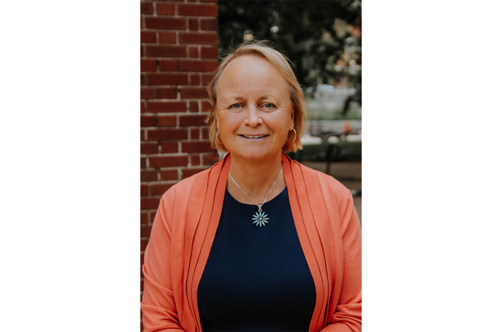 Photo of Sue Roberts A person with short light-colored hair is standing outdoors in front of a brick wall, wearing a dark top with a coral cardigan and a silver necklace featuring a circular pendant.