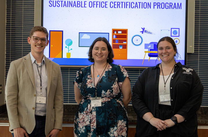 photo from Sustainable Office Certification Program Three people stand indoors in front of a large screen displaying the text “Sustainable Office Certification Program.” The screen shows colorful icons related to sustainability, including plants, a recycling symbol, and office items.