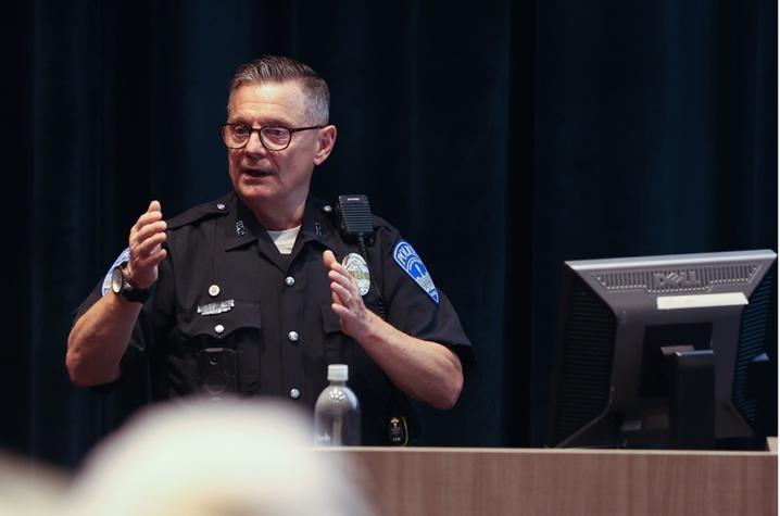 Officer Saylor teaching a staff safety course in a theater on campus. He's in uniform, talking at a lectern with his hands in the air.