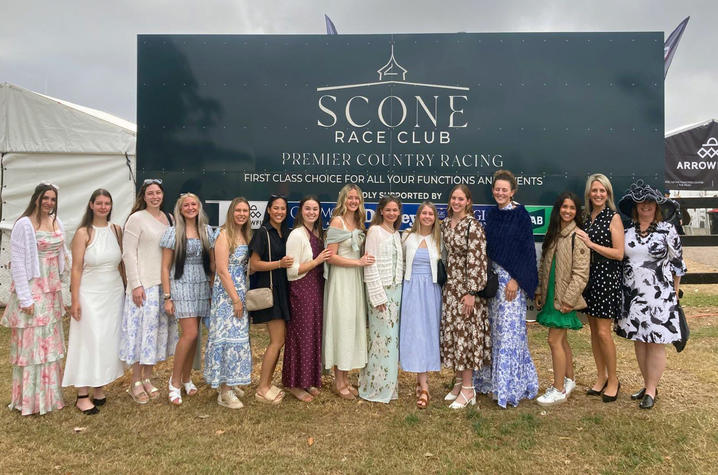 A group of people dressed in colorful dresses, skirts and race-day attire poses for a photo in front of a large sign reading “Scone Race Club” at an outdoor horse racing venue. 