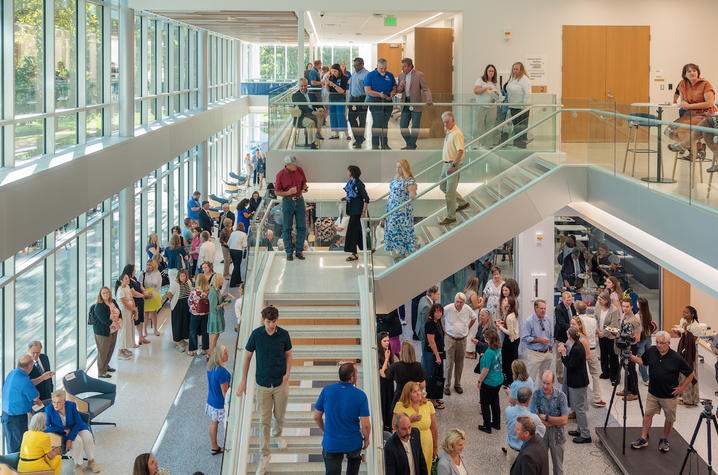 A spacious, multi-story atrium is filled with people attending a gathering inside a modern building. Large windows line the left side of the space, allowing abundant natural light to stream in and illuminate the interior.