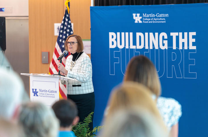 A person stands at a podium speaking to an audience during an indoor event. Behind the speaker, a large blue backdrop displays the words “BUILDING THE FUTURE” in large white letters.