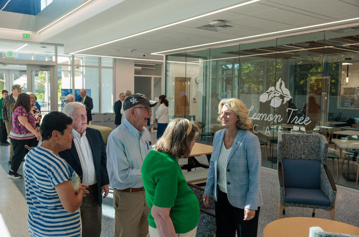 Several people gather inside a bright lobby or reception area with floor-to-ceiling glass walls and abundant natural light.