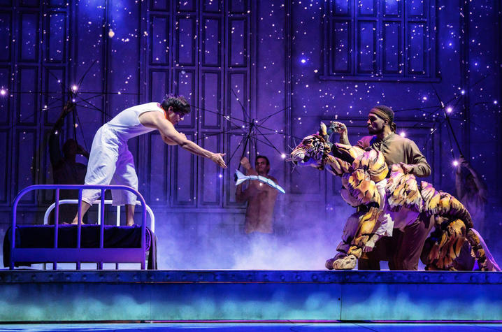 An actor standing on a bed feeding a fish to a Bengal tiger puppet.