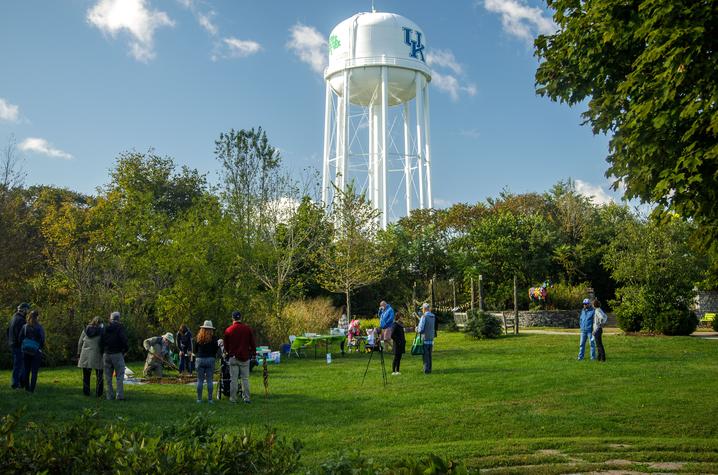 Arboretum during Arbor Day A group of people gathered on a grassy area surrounded by trees, participating in an outdoor activity under a clear blue sky. In the background, a tall white water tower with the letters “UK” painted in blue is visible above the treetops.