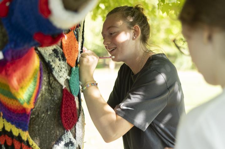 students with the Living Learning Program at The Arboretum Person attaching colorful crocheted or knitted pieces to a tree trunk outdoors. The yarn pieces include bright colors like red, blue, yellow, and green, forming a patchwork design. The background shows green foliage and sunlight filtering through trees