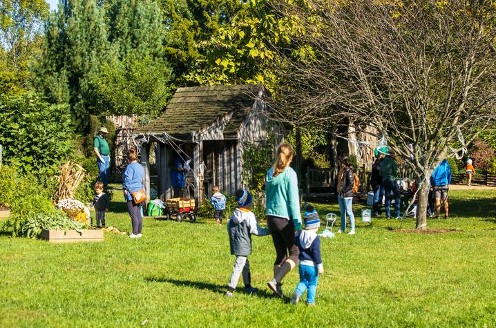 Kentucky Children’s Garden People gathered outdoors near small rustic wooden cabins surrounded by greenery. Some individuals are standing and talking, while others are walking across the grassy area with children. Raised garden beds and trees are visible in the foreground.