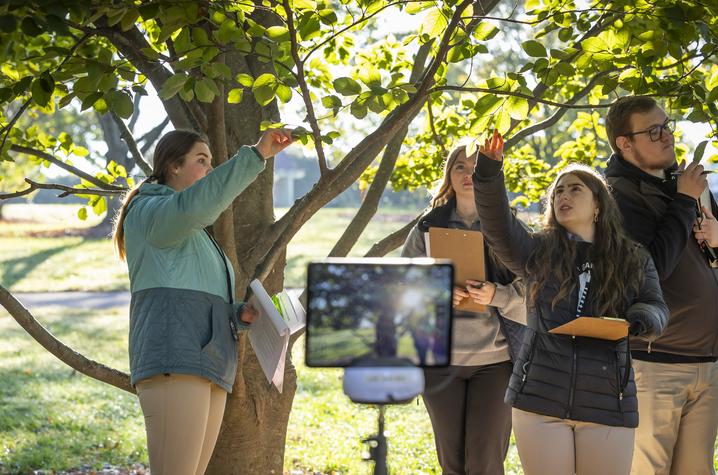 Teaching simulation at The Arboretum Several people standing under a tree, holding clipboards and papers, appearing to observe or record something outdoors. A tablet on a tripod is positioned in the foreground, facing the group.