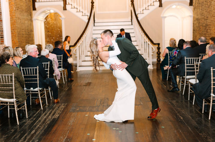 Photo of Carly and Nick Lovely at their wedding Two people in formal wedding attire share a kiss while one person dips the other on a wooden aisle inside an elegant indoor venue, with seated guests on both sides and a curved staircase in the background.