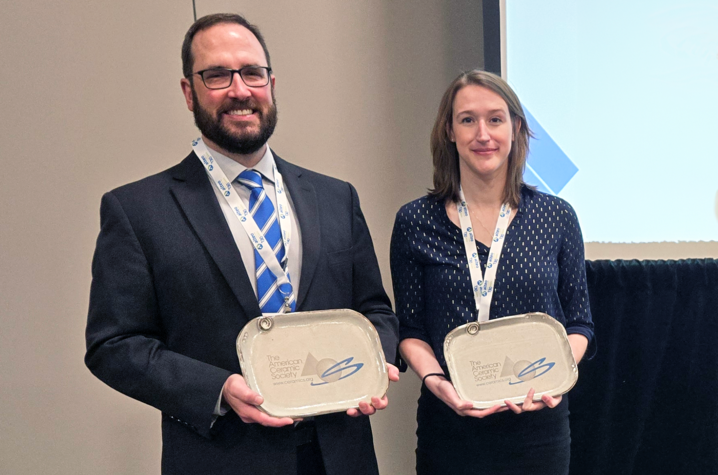 Bob Jewell and Tristana Duvallet standing in a room in front of a screen on which a presentation is being projected. They are each holding awards. 