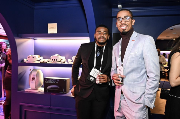 Photo of Cameron Brown Two men wearing conference badges pose and smile at a networking event inside a modern, blue-lit venue. One wears a black suit and holds a drink, while the other wears a light gray suit with a dark turtleneck and glasses.