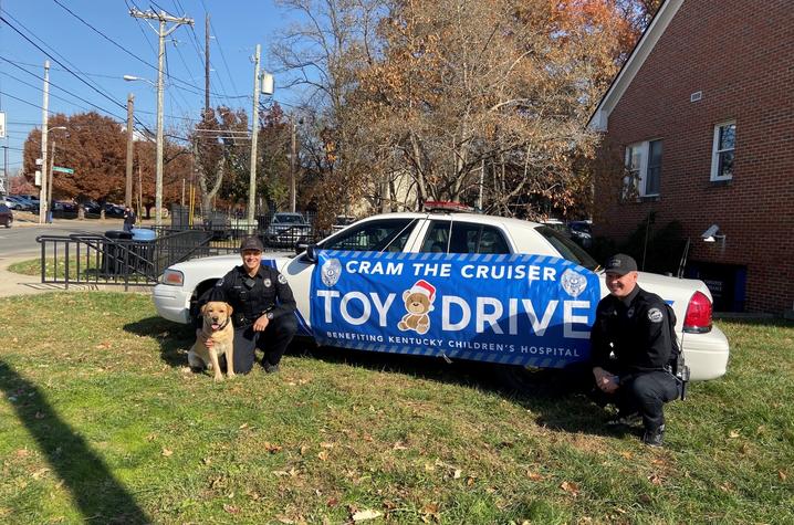 Photo of Officer Michael Culver, K-9 Hudson and Officer Ryan Johnson in front of the cruiser with a banner that says "Cram the Cruiser" toy drive.