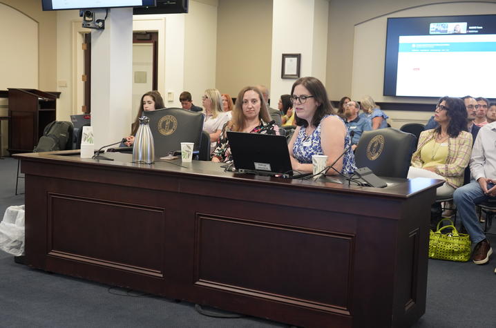 People seated behind a long wooden dais use laptops and documents during a formal meeting, with a presentation screen visible in the background.
