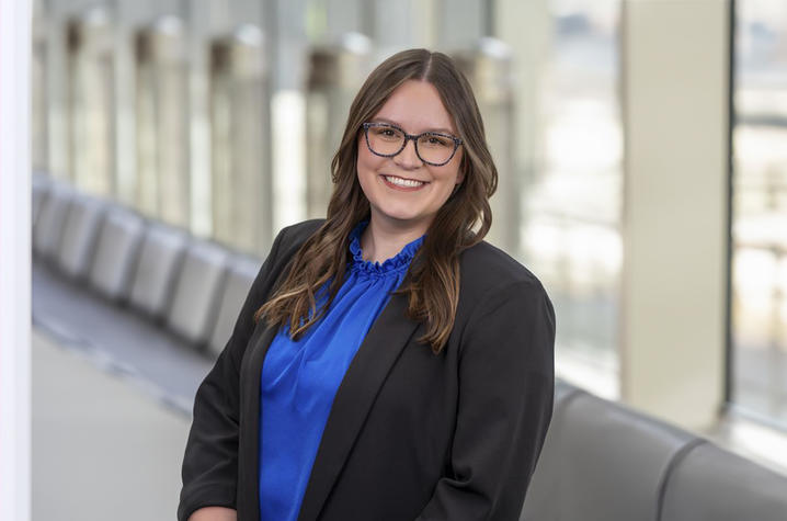 Class president Ramey Hensley of Fort Thomas, Kentucky, matched with the UKCOP/PrimaryPlus Community-Based Residency. A woman wearing glasses, a blue blouse, and a black blazer smiles while standing in a bright hallway with large windows. The background is softly blurred, emphasizing her professional portrait.
